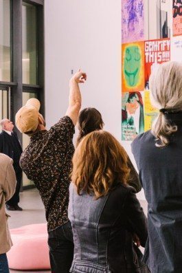 The photo shows six people from behind in front of the poster wall. A young man with a cap is pointing at one of the posters.