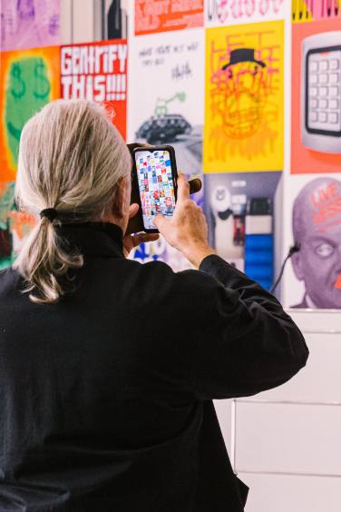 The picture shows a man from behind. He has long grey hair tied back in a plait and is wearing a black overall. He is taking a photo of the poster wall.