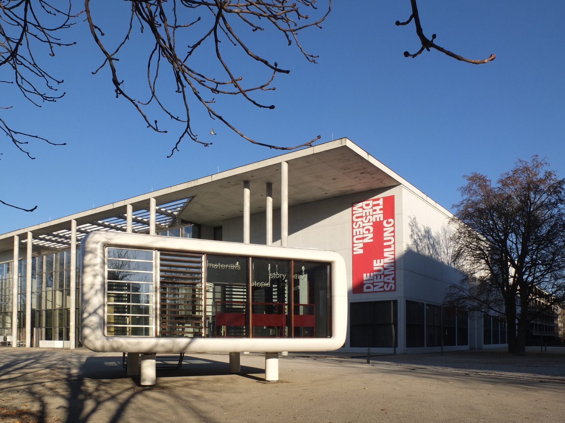 Exterior shot with a view of the Loftcube and the entrance area of the Pinakothek der Moderne. The Loftcube is a cuboid with rounded corners. The walls are made of glass, the cladding is white. It stands on four cylindrical legs.