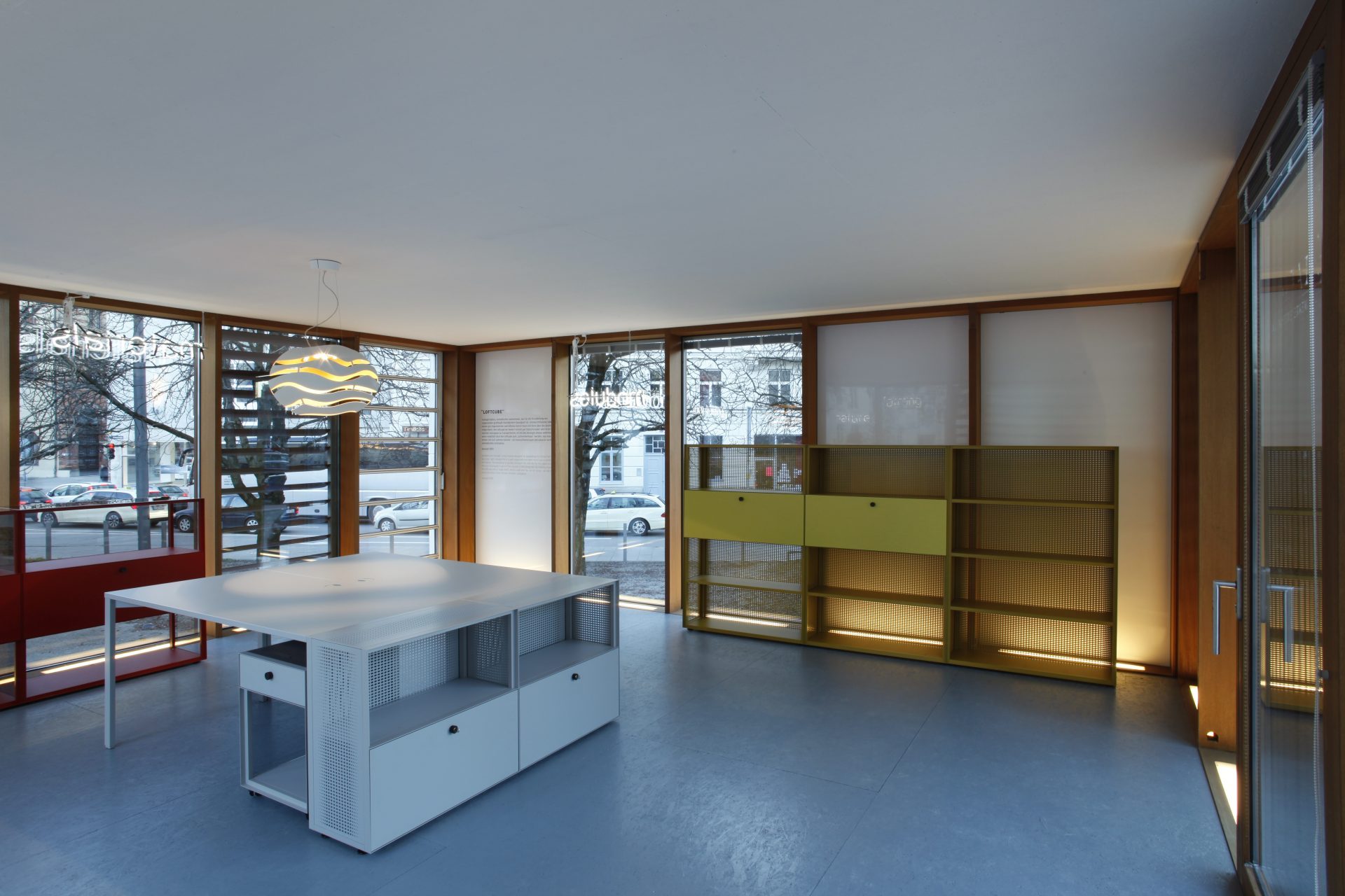 Photo of an interior. The glass walls are lined with yellow and red shelves with drawers and open compartments. In the centre of the room is a white table with storage compartments on one side.