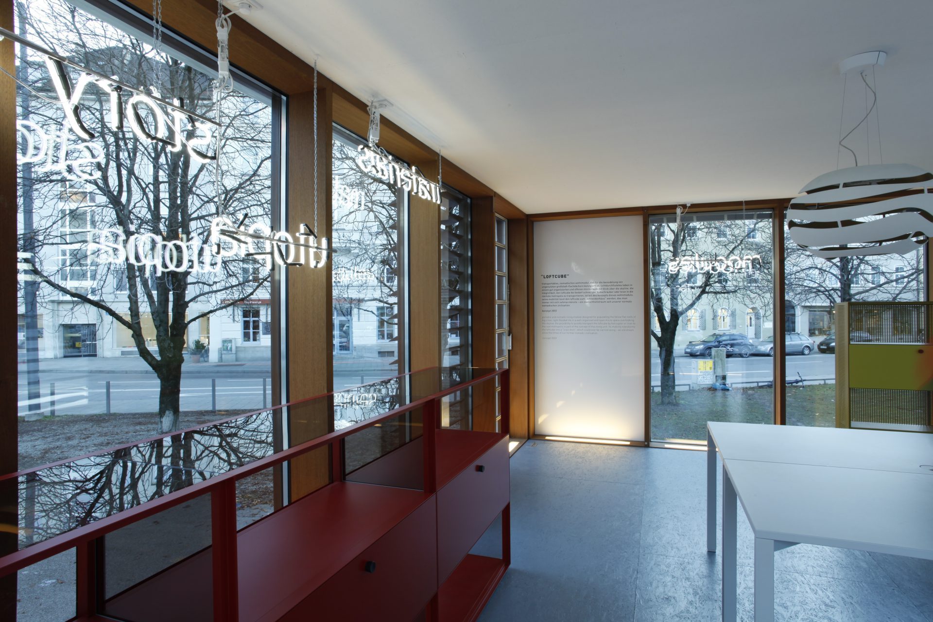 Interior view. Detailed view of a glass wall with illuminated lettering and a red shelf. To the right is a white table at the edge of the picture. An exhibition text is printed on the wall opposite.