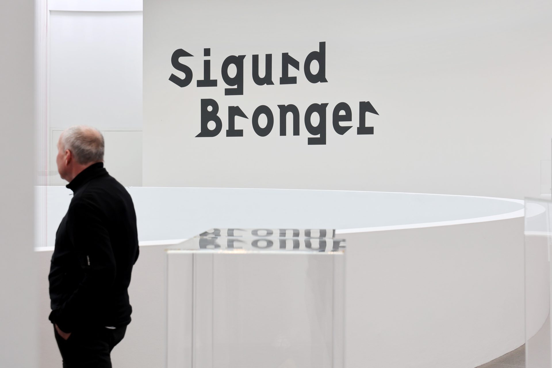 A man is standing in the Sigurd Bronger exhibition. He is wearing a black turtleneck and black trousers. He is depicted from the side and moves through the room. His face is unrecognisable. He has brown hair and is bald. The title of the exhibition can be seen on the wall in the background.