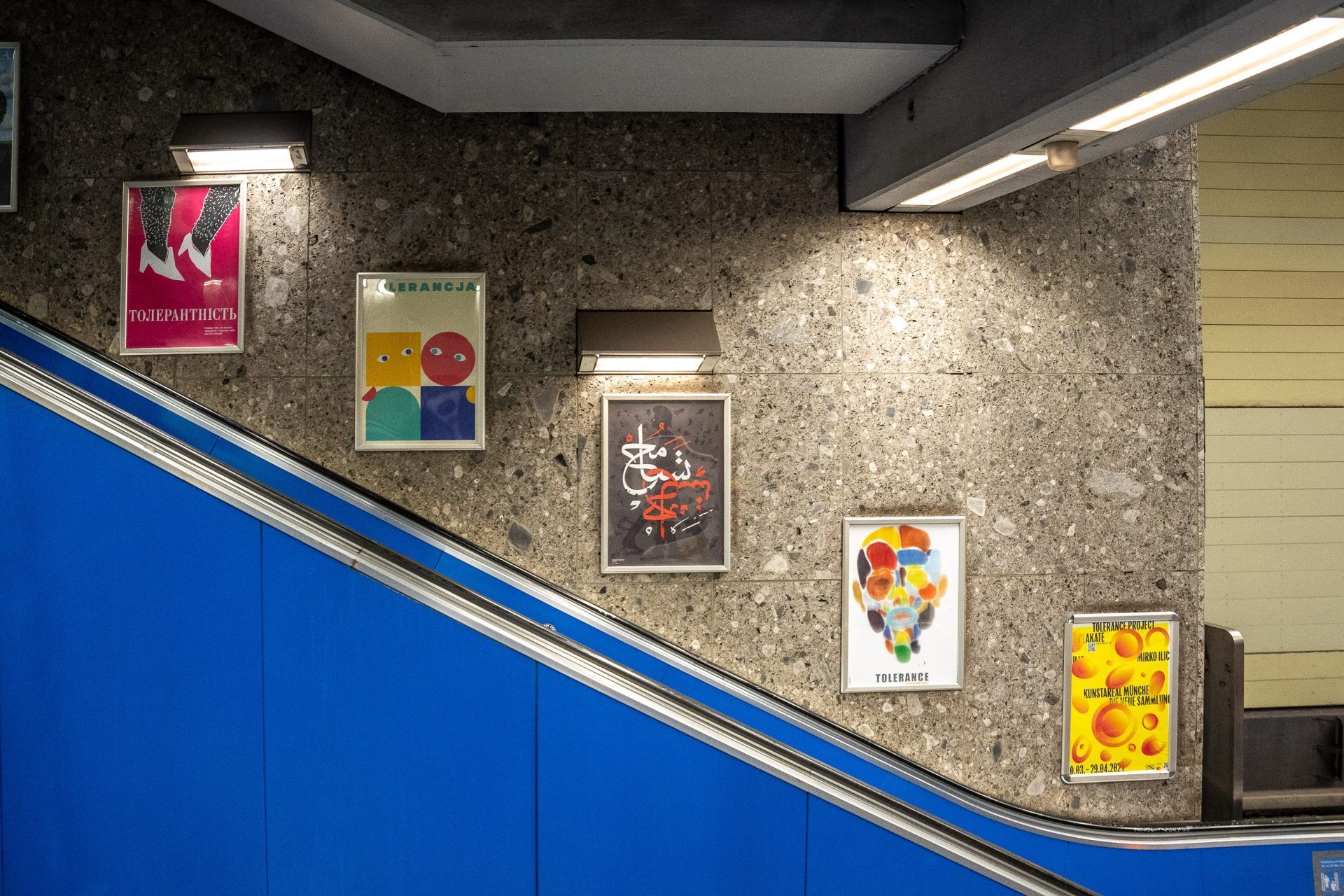 5 different posters from the exhibition hang above a blue escalator.