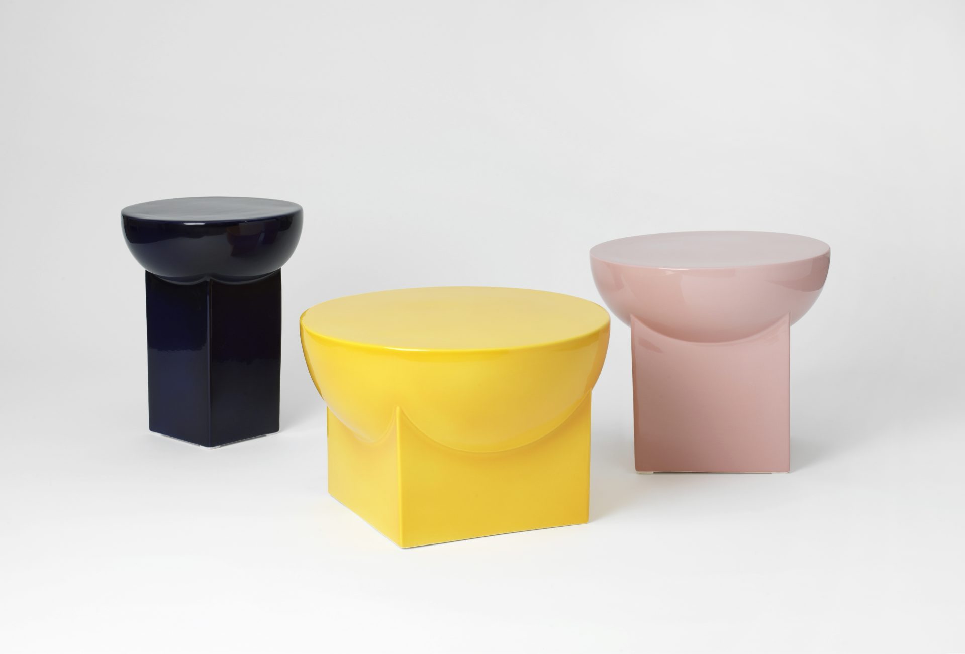 Three modern, sculptural side tables with a glossy surface in the colours dark blue, yellow and pink, arranged on a white background.