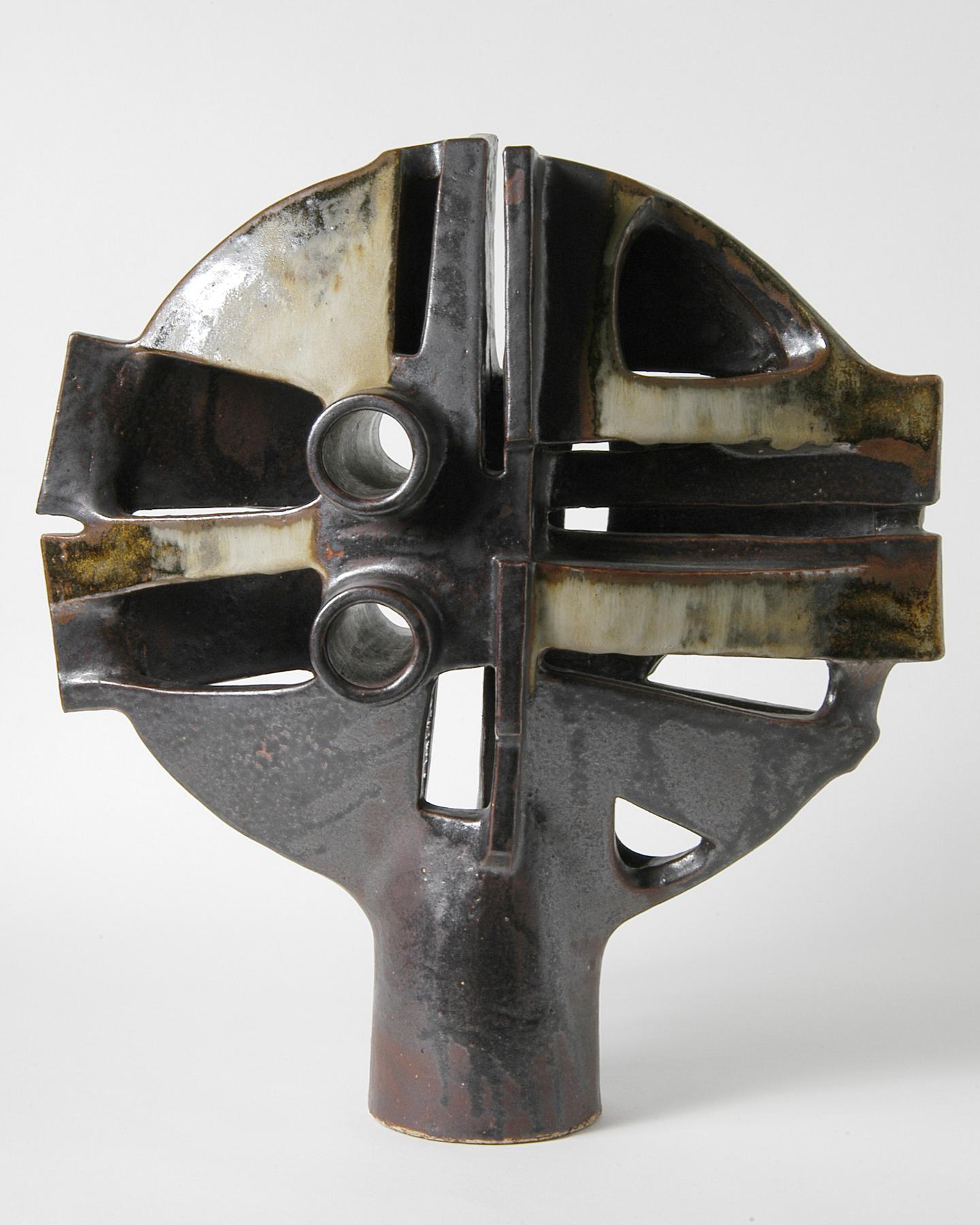 Sculptural ceramic with a symmetrical, abstract shape, dark glaze and light-coloured accents, pierced with round and rectangular openings.