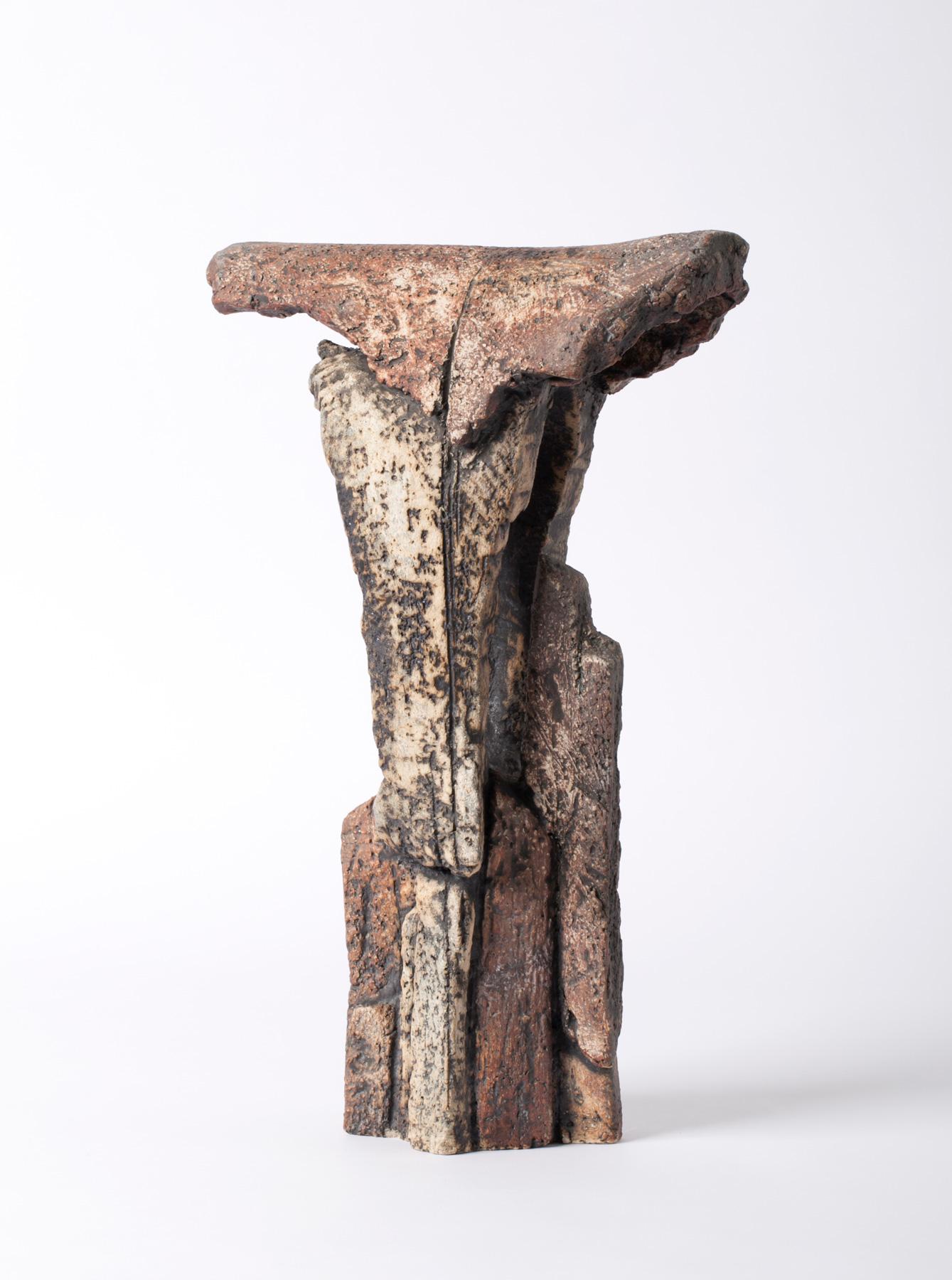 Skulpturale Keramik mit verwitterter Oberfläche in Erdtönen. Die Form erinnert an eine erodierte Steinsäule mit einer breiten, unregelmäßigen Oberseite und tiefer reliefartiger Struktur.