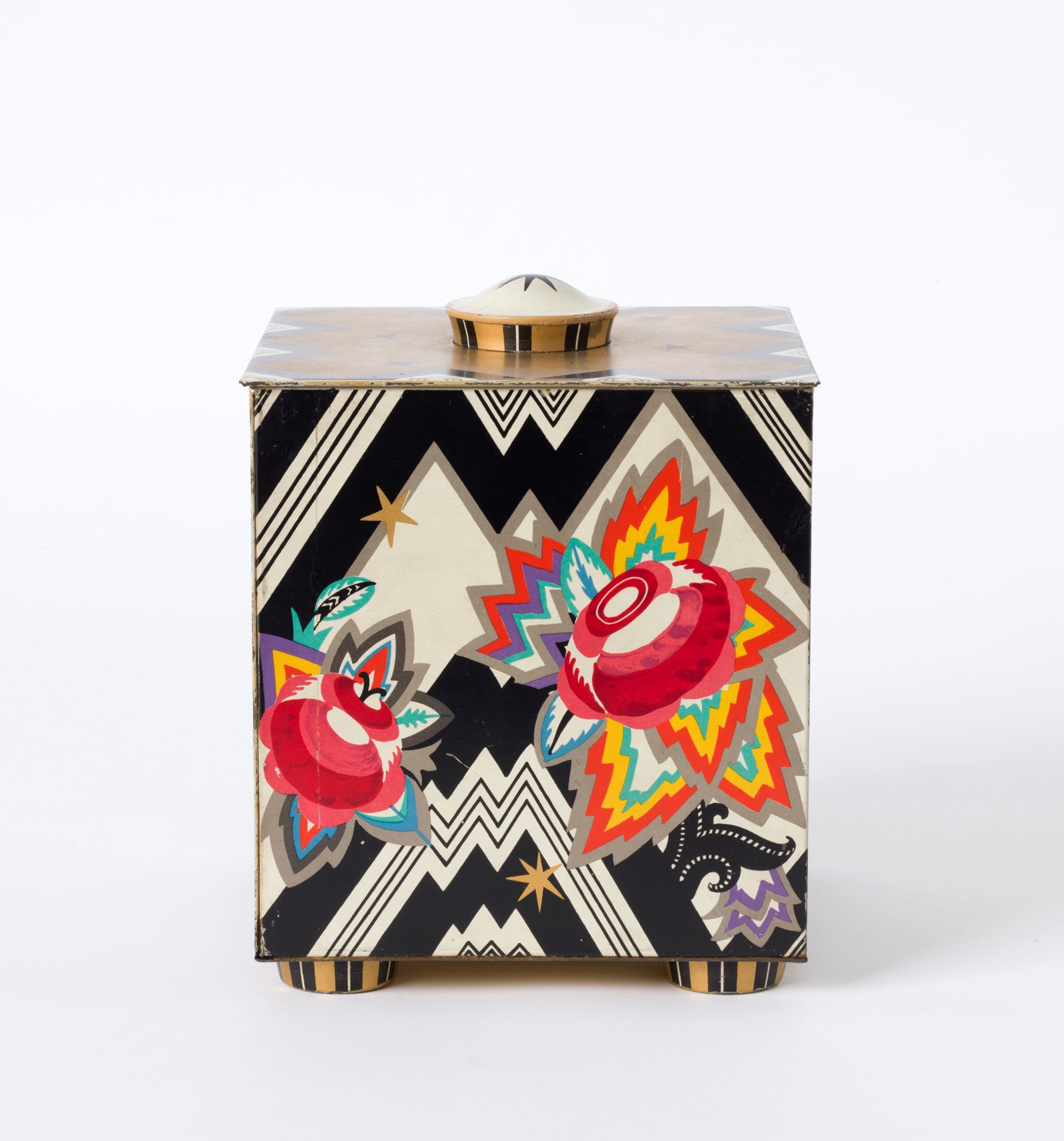 Photo colorful can. The box is rectangular and stands on four small, round feet. There is a round knob on the lid at the top center. The pattern on the can is jagged black and white and there are flowers in bright colors on it.