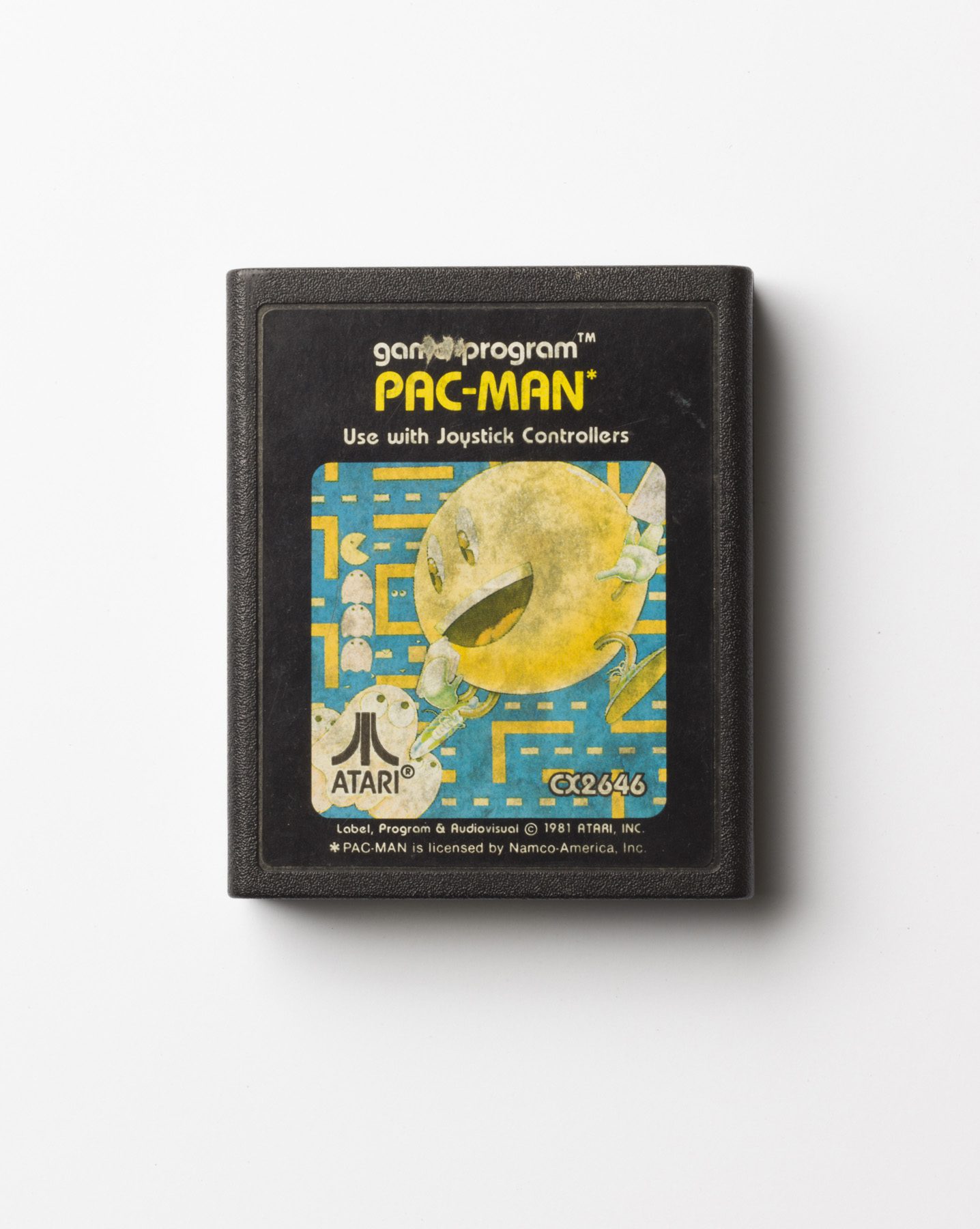 Black Atari game cartridge with the title ‘PAC-MAN’ in yellow lettering. The label shows an illustration of Pac-Man, a yellow ball with arms and legs, running through a maze, pursued by ghosts. 