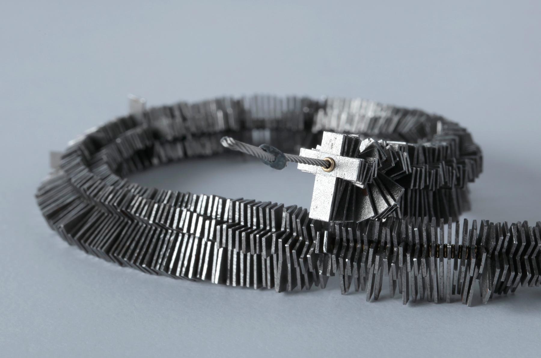 Necklace made of stacked, metallic cross elements with a textured clasp, against a grey background.