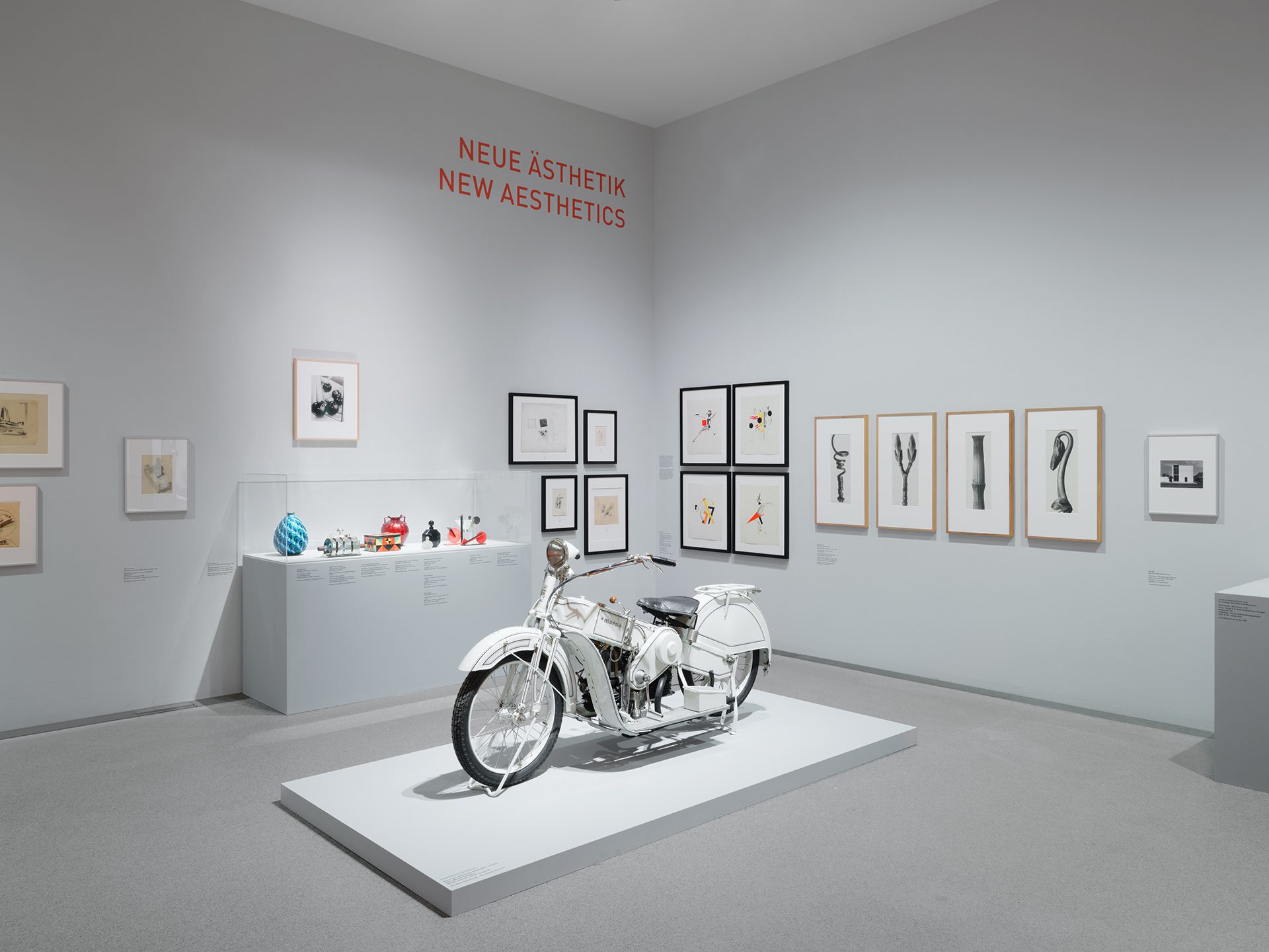 You can see the exhibition installation "4 Museums – 1 Modernism." In the foreground is a motorcycle, behind it is a display case with objects. Framed pictures from the exhibition hang on the walls.