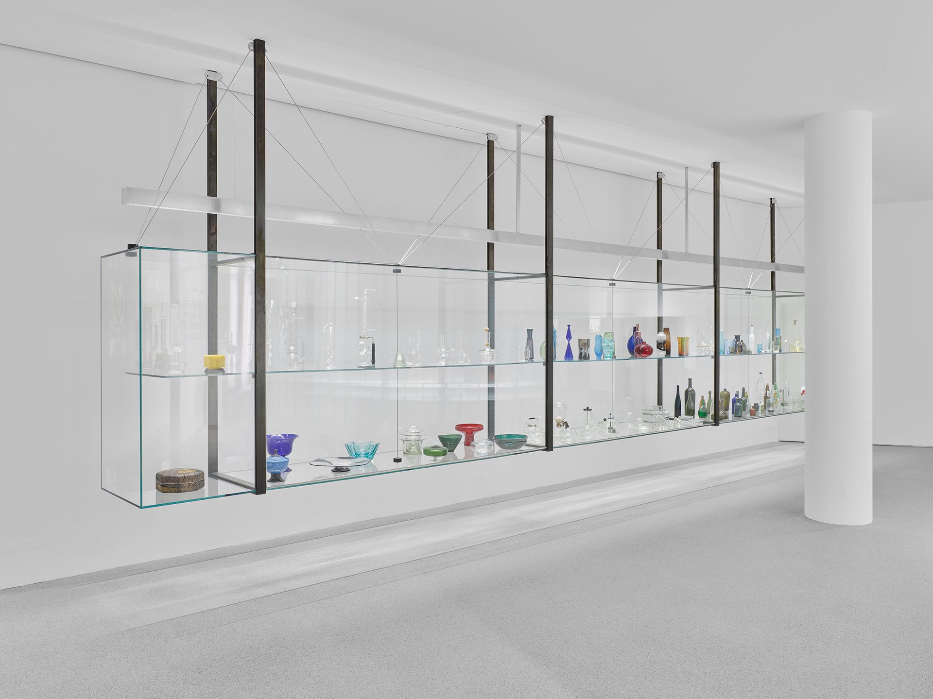 Man sieht die Ausstellungsinstallation "Die Farbe von Glas" eine an der Decke befestigte Glasvitrine in der auf zwei Ebenen verschiedene Objekte aus Glas gezeigt werden.