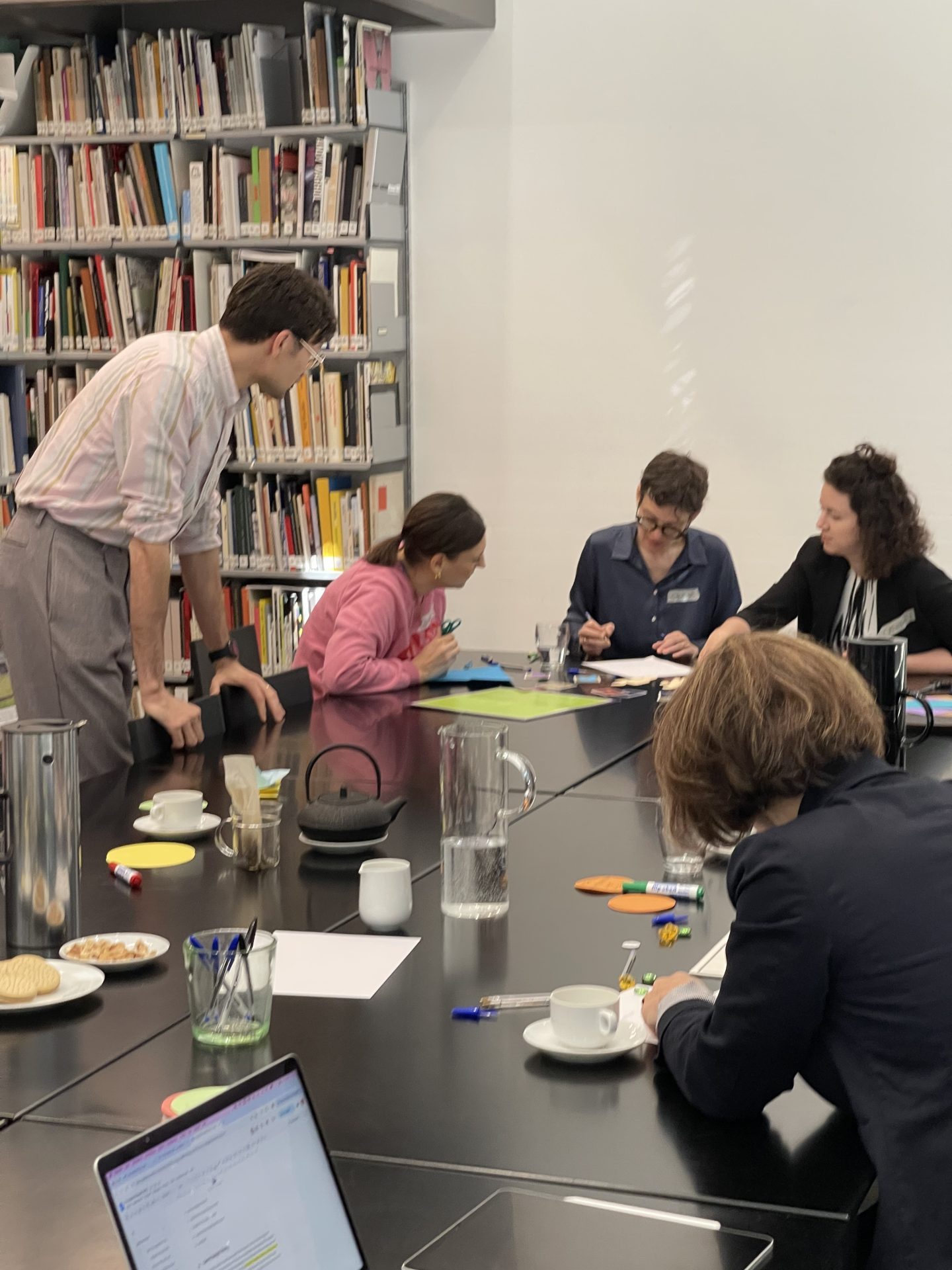 Eine Gruppe von Menschen sitzt an einem schwarzen Tisch mit Laptop, Wasserkaraffe und Papieren. Links hinter Ihnen ist ein Bücherregal.