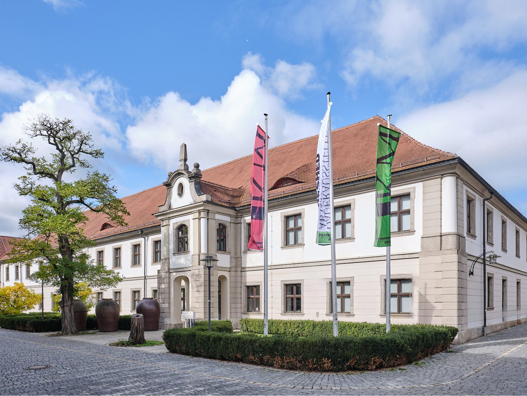 Historisches Gebäude mit Fahnen, alten Bäumen und einem gepflasterten Weg unter blauem Himmel.