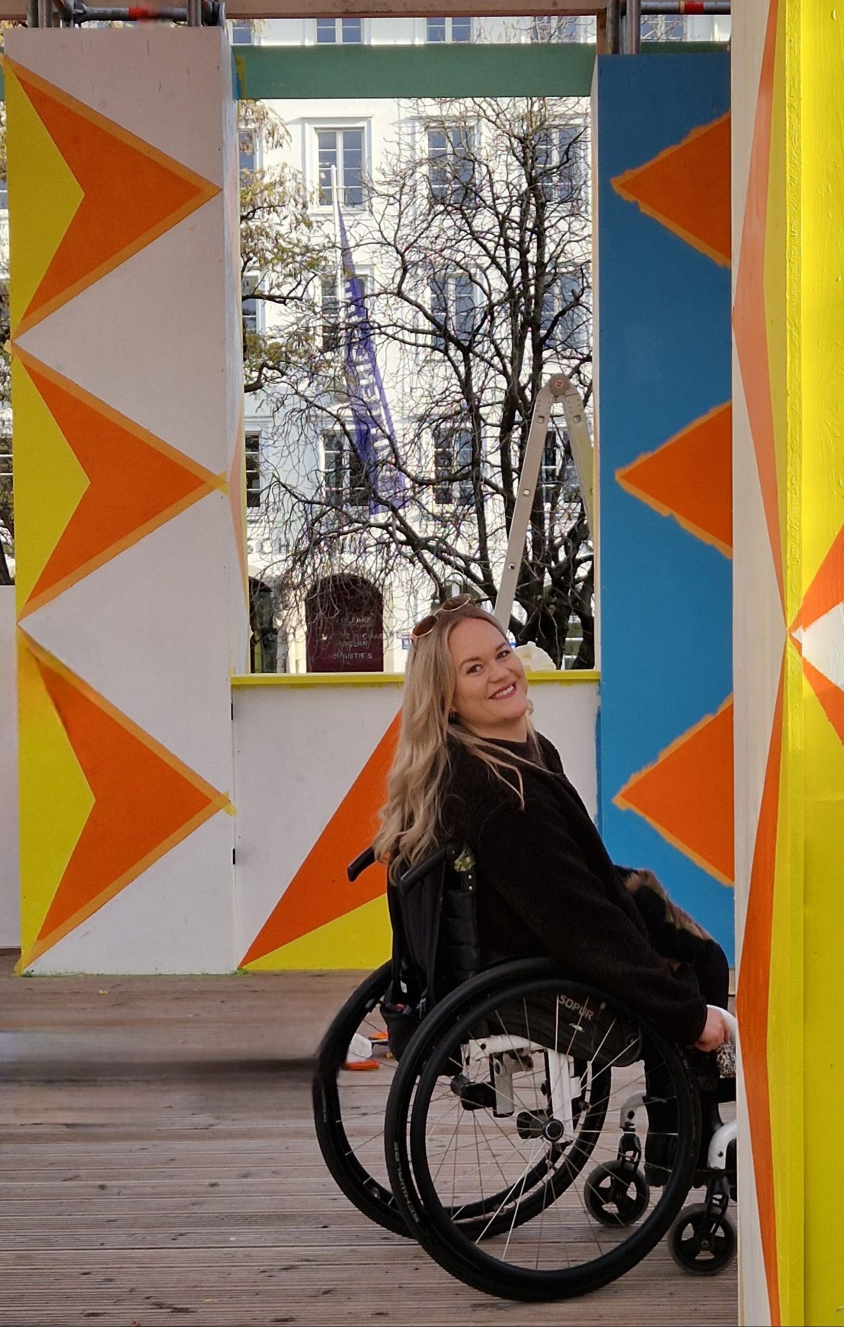Blonde Frau im Rollstuhl in bunter Architektur.