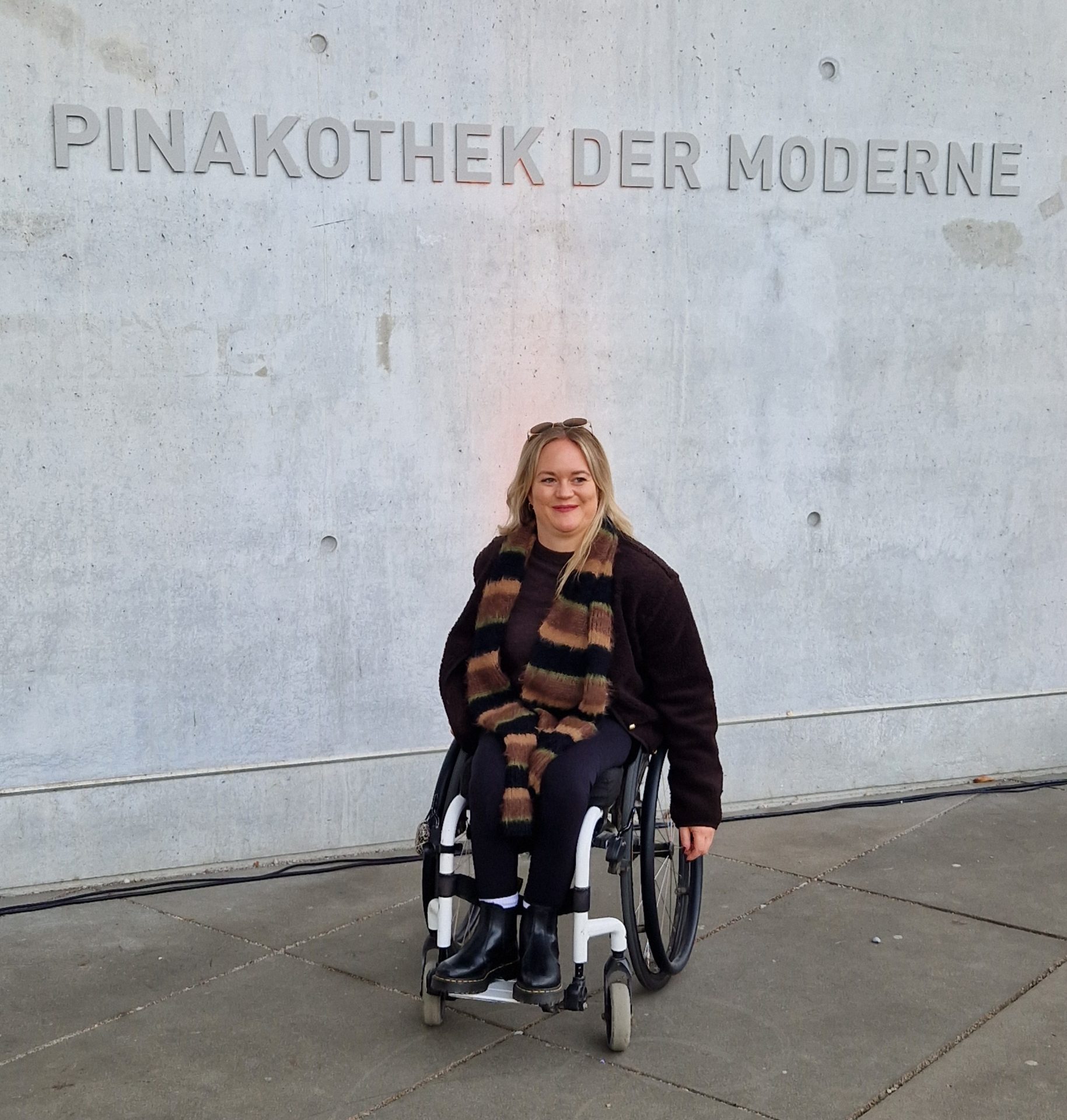 Eine Frau mit blonden Haaren und im Rollstuhl vor einer grauen Wand. An der Wand steht über ihr Pinakothek der Moderne.