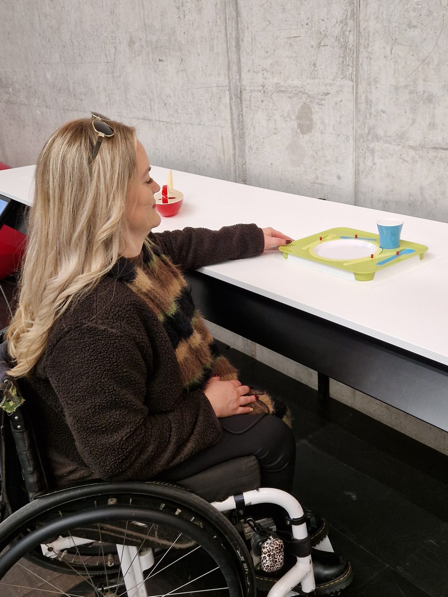 Frau im Rollstuhl an weißem Tasttisch berührt mit ausgestrecktem Arm einen Kinderteller mit Becher.