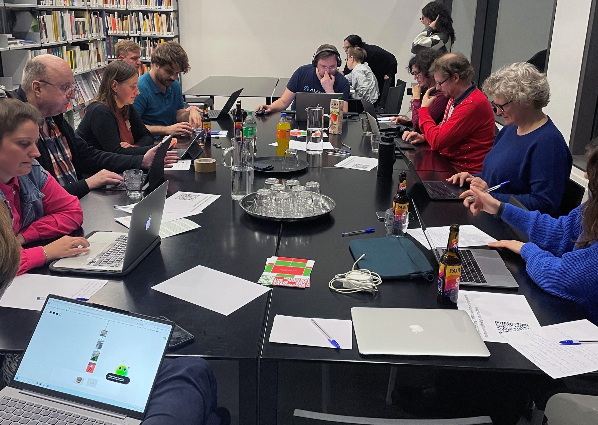 Eine Gruppe von Menschen sitzt an einem großen schwarzen Tisch mit Laptops und Tablets. In der Mitte stehen Getränke.