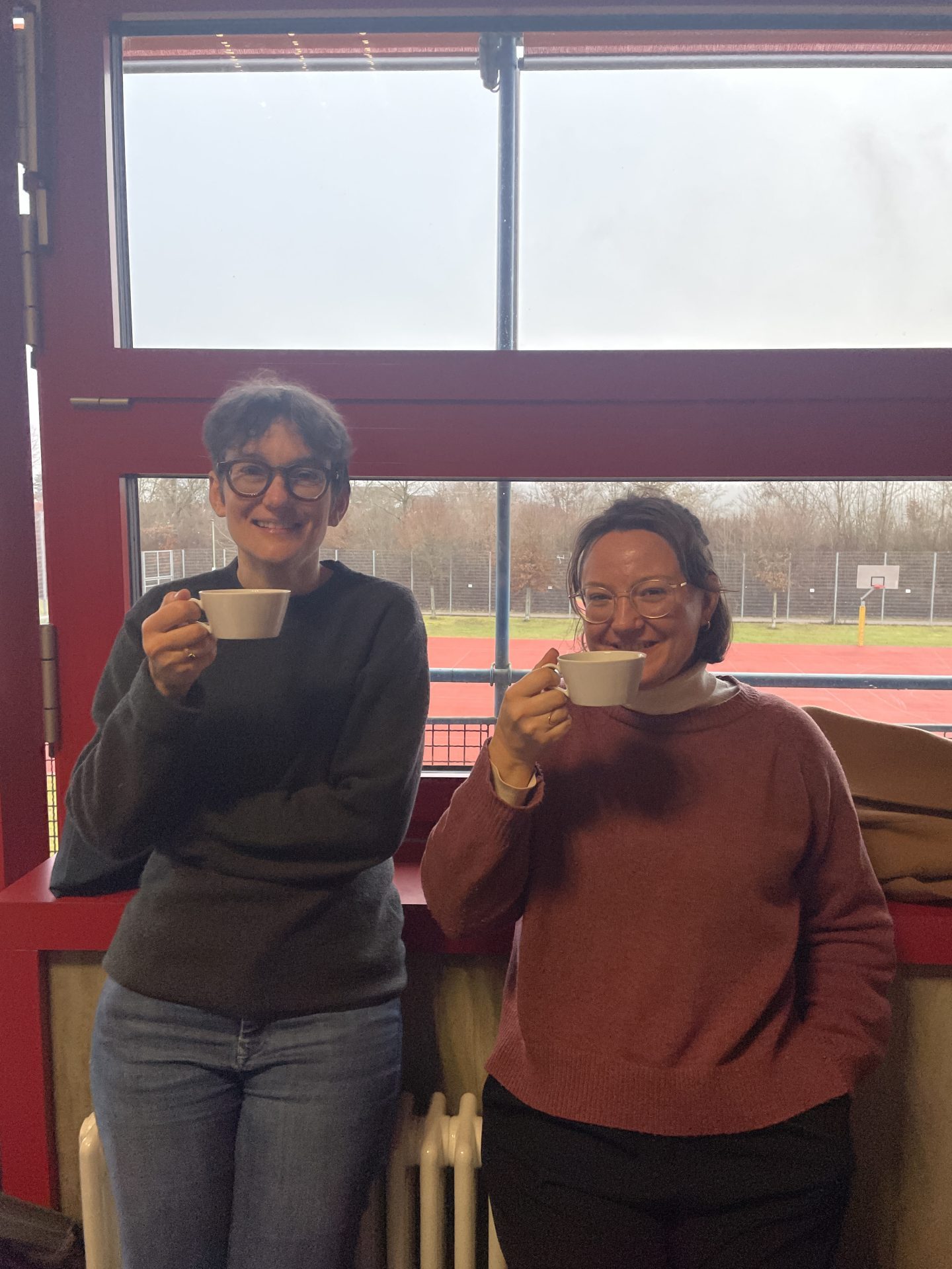 Zwei Frauen vor einer Fensterfront lehnen sich an eine Heizung und trinken Tee.