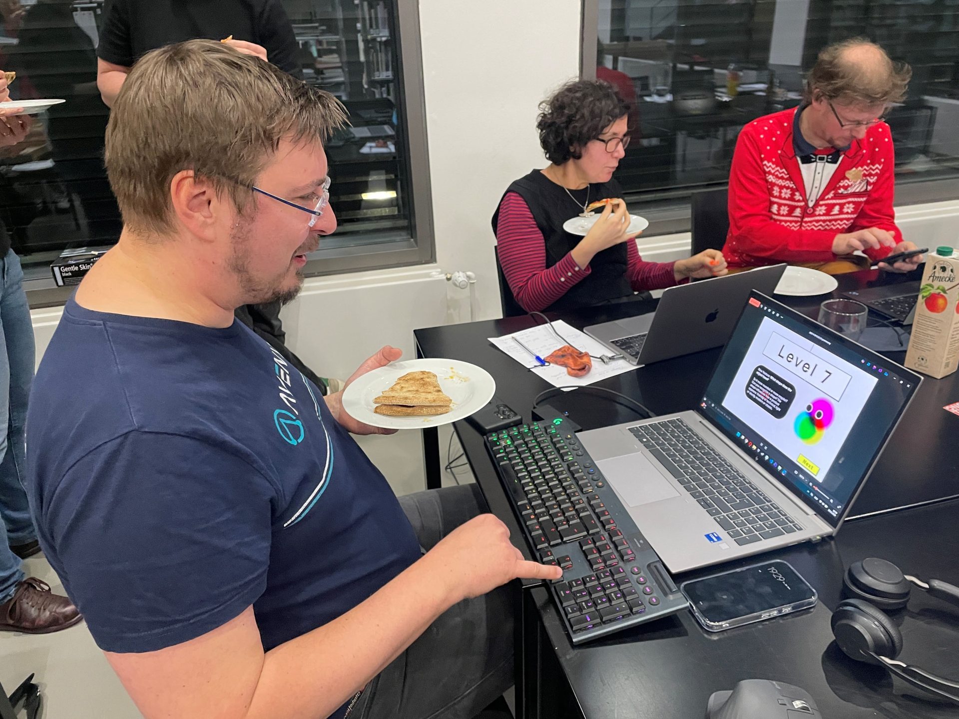 Foto Mann mit Pizza auf Teller in einer Hand arbeitet mit der anderen am Laptop. Im Hintergrund zwei weitere Personen mit Essen und Laptops.