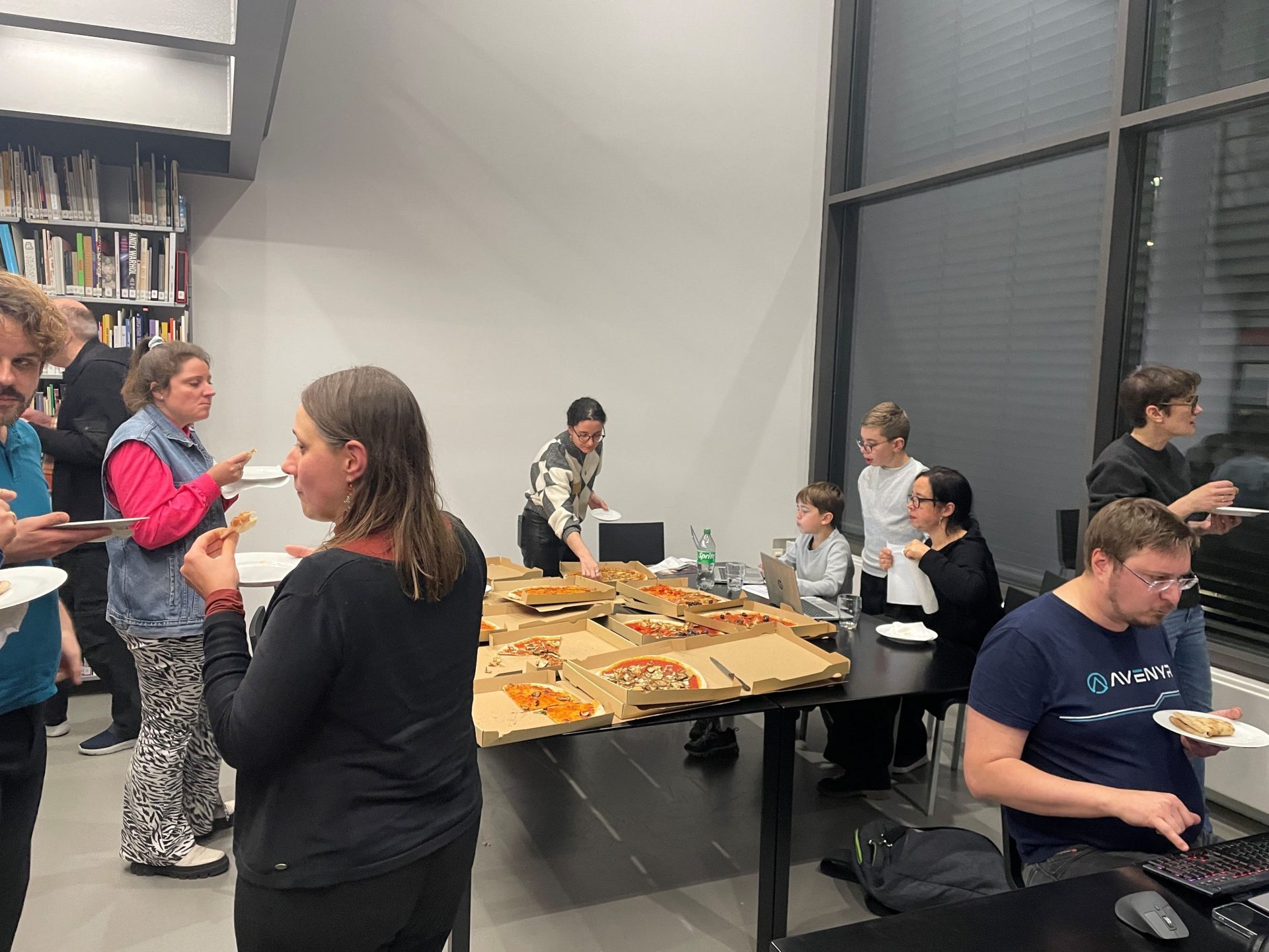 In der hinteren Ecke des Raums sind große Pizzen auf einem Tisch ausgebreitet. Darum stehen mehrere Personen mit Pizzastücken in der Hand und unterhalten sich.