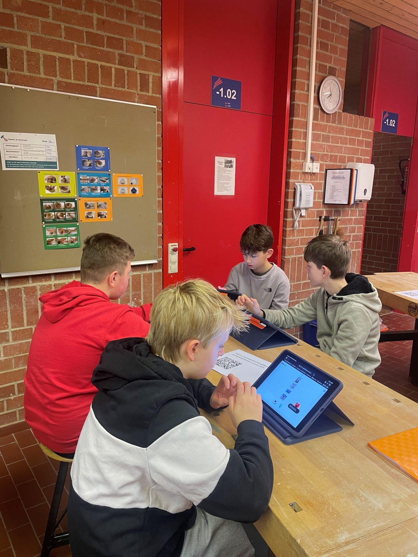 Innenaufnahme Klassenzimmer, Werkraum. Vier Jungs sitzen um eine Werkbank, auf der sie konzentriert mit ihren Tablets ein Game spielen.