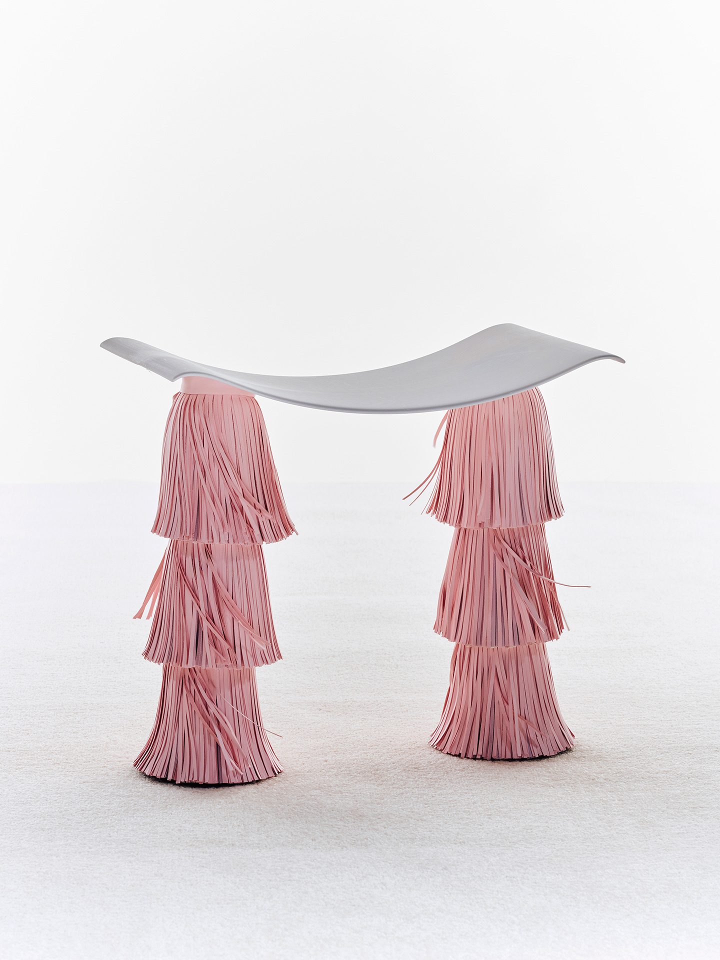 Hocker mit nach unten gewölbter grauer Sitzfläche und zwei Füßen mit rosa Fransen.