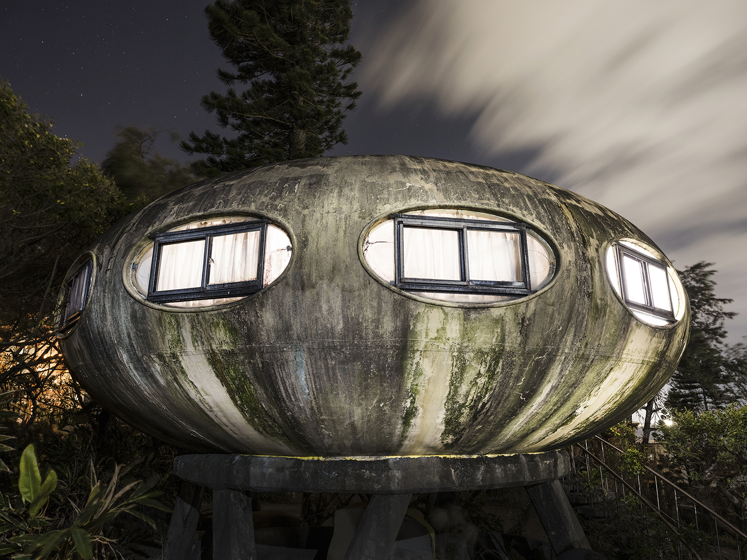 A heavily weathered exterior view of a replica of the Futuro plastic house, featuring a concrete plinth and steps.