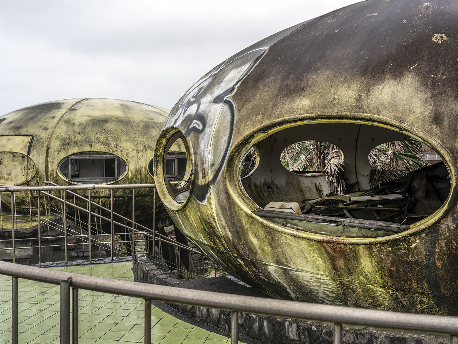 A heavily weathered exterior view of a replica of two Futuro plastic houses, featuring a green-tiled terrace and metal handrails.